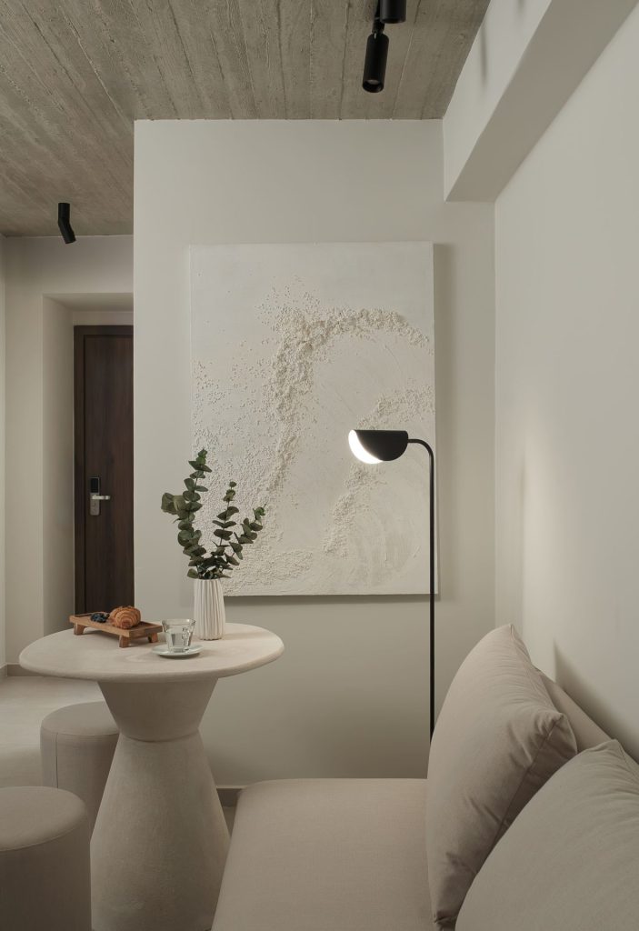 Minimal Japandi καθιστικό με στρογγυλό τραπέζι, μπεζ καναπέ, εμφανές μπετόν στην οροφή και μοντέρνο έργο τέχνης στον τοίχο, Nerea Suites.